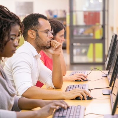 Multiracial group of students training in computer class Multiracial group of students training in computer class. Line of man and women in casual sitting at table, using desktops, typing, looking at monitor. Training center concept
